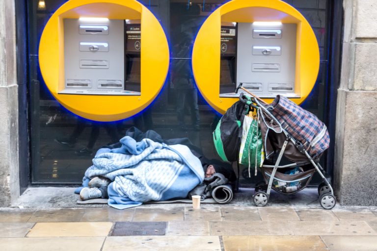 Hombre sin techo cubierto con mantas, que duerme bajo unos cajeros automáticos con un cochecito y sus pertenencias