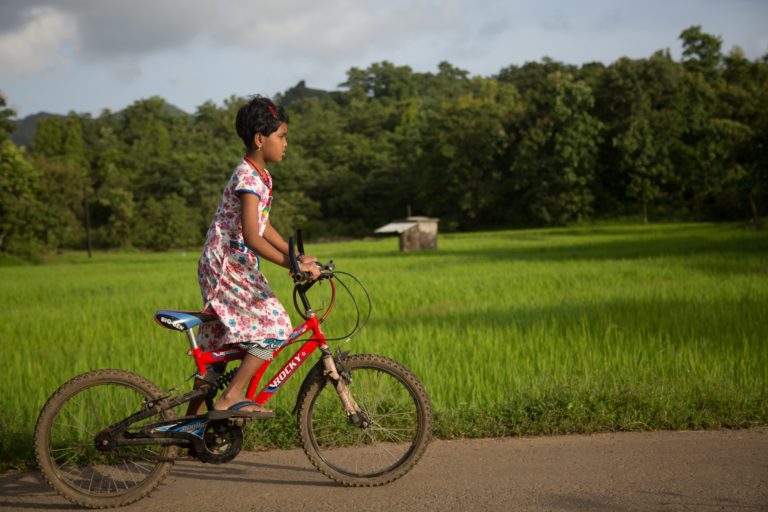 Una niña en una bicicleta roja en la India rodeada de verdes campos